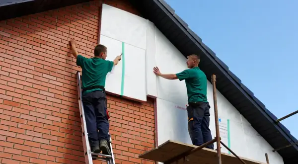 Deux ouvriers réalisent l'isolation par l'extérieur soi même d'une maison en briques, fixant des panneaux isolants blancs sur le pignon.