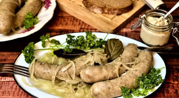 Saure Bratwurst mit Zwiebeln und Petersilie auf einem Teller, dazu Brot und ein Glas Senf.