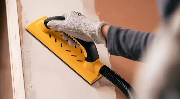 Mano enguantada usando una lijadora amarilla para lijar paredes, preparando la superficie para pintar.