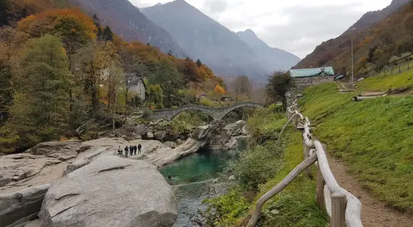 Sentiero sul monte Valdo, con un ponte ad arco in pietra, un fiume turchese e montagne sullo sfondo.