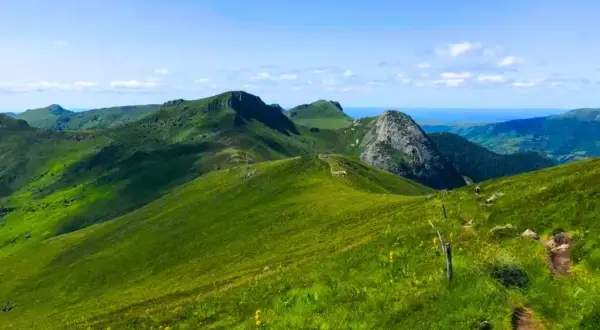 Paysage de montagnes verdoyantes sous un ciel bleu, idéal pour les plus belles randonnées dans le Cantal. Un sentier serpente sur la colline.