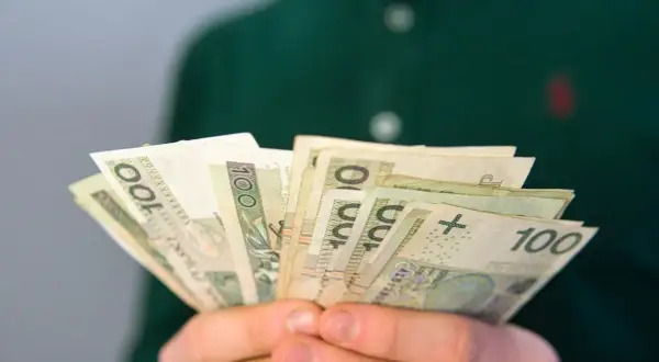 Dłonie trzymające plik banknotów 100 zł. Zastanawiasz się, ile wynosi renta rodzinna dla dziecka?