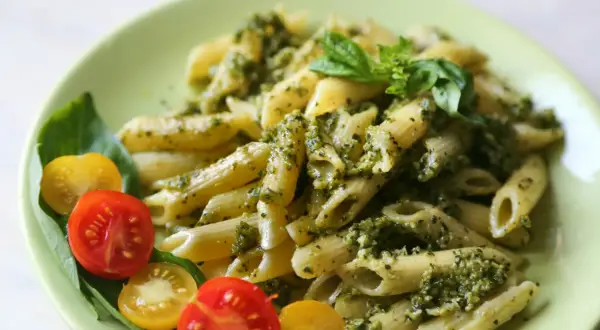 Makaron penne z sosem pesto, pomidorkami koktajlowymi i bazylią na zielonym talerzu.