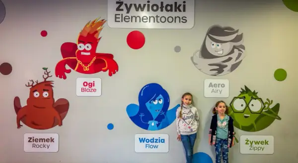 Dwie dziewczynki pozują na tle ściany z postaciami z bajki "Żywiołaki Elementoons". Widać Ogi Blaze, Ziemka Rocky, Wodzię Flow, Aero Airy i Żywka Zippy.