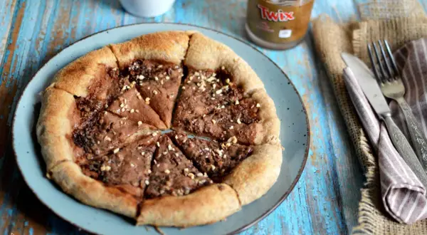 Pizza al cioccolato con crema Twix e nocciole, cosparsa di granella croccante. Un dolce goloso pronto da gustare.