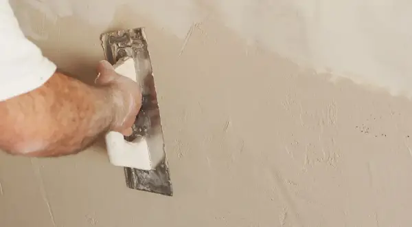 Mano che stende l'intonaco rasato con una spatola, creando una finitura liscia e uniforme su una parete.