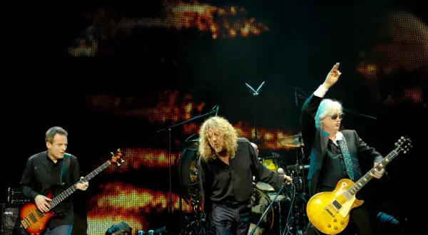 Trzech muzyków na scenie podczas koncertu Led Zeppelin. John Paul Jones gra na basie, Robert Plant śpiewa, a Jimmy Page z uniesioną ręką gra na gitarze.