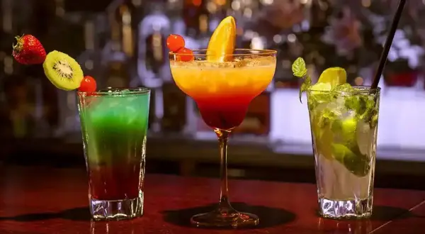 Trzy kolorowe drinki: zielono-czerwony z owocami, pomarańczowo-czerwony w kieliszku oraz zielony mojito.