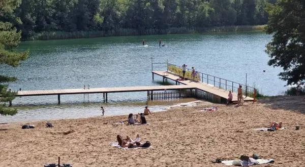 Słoneczna borówno plaża nad jeziorem, ludzie odpoczywają na piasku i w wodzie, niektórzy pływają na desce SUP.