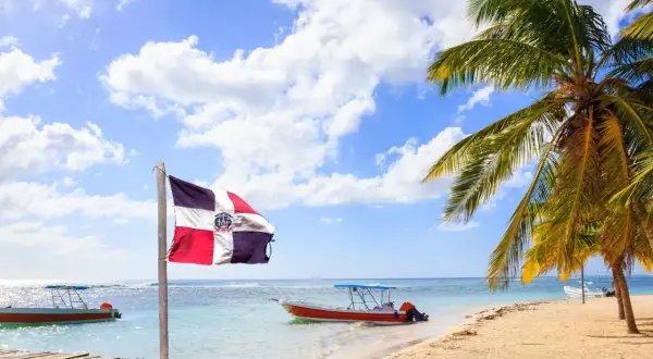 Dominikanische Flagge weht am Strand. Boote im türkisfarbenen Meer. Palmen. Hier ist es im Januar warm.