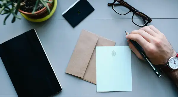 Ręka pisze o uciążliwym sąsiedzie. Na biurku notatnik, okulary, koperta i papeteria.