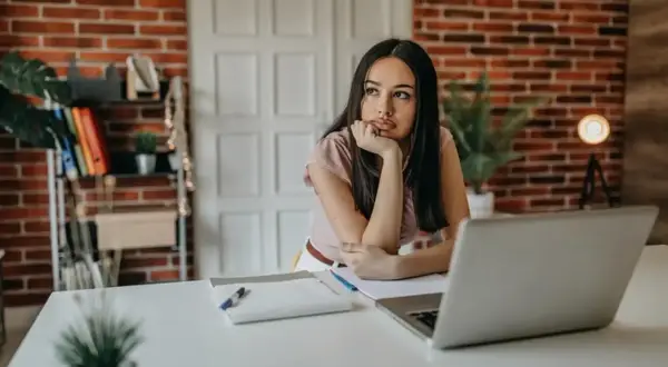 Młoda kobieta siedzi przy biurku, zamyślona, przeżywa kryzys 30 latka. Laptop i notatki leżą przed nią.