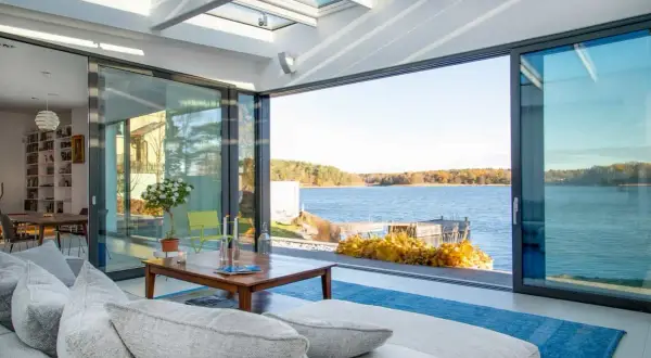Soggiorno luminoso con vista lago, divano grigio e tavolino in legno. L'ampia vetrata e il soffitto trasparente creano un perfetto stile coastal.