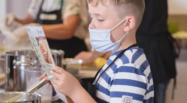 Junge mit Maske liest Rezept, während andere kinder kochen selbst. Küchenutensilien und Töpfe sind zu sehen.