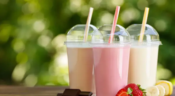Trzy kolorowe szejki w plastikowych kubkach z rurkami, obok truskawki, banany i kawałki czekolady.