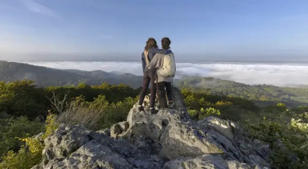 Zwei Freunde genießen den Ausblick beim Wandern an der Sieg. Nebel liegt im Tal, die Sonne scheint auf die Berge.
