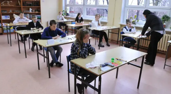 Uczniowie siedzą w ławkach podczas egzaminu szóstoklasisty. Jedna z uczennic zakrywa twarz dłońmi, widać napięcie.