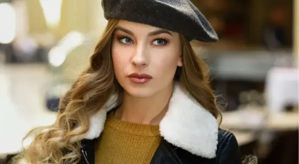 Elegancka kobieta w grafitowym beretu, z burzą blond loków i w czarnej ramonesce. Dowiedz się, jak nosić beret damski, by wyglądać stylowo.