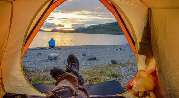 Blick aus dem Zelt auf einen See bei Sonnenuntergang. Eine Person sitzt am Ufer. Ja, darf man in Schweden wildcampen!