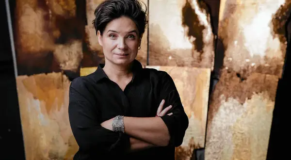 Agnieszka Smoczyńska, reżyserka o krótkich, ciemnych włosach, w czarnej koszuli, z rękami skrzyżowanymi na piersi, na tle abstrakcyjnych, złotobrązowych płócien.