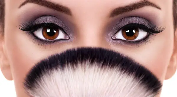 Occhi marroni intensi, evidenziati da un trucco smokey viola e ciglia lunghe, sembrano ancora più grandi e sporgenti grazie a un pennello da trucco sfocato.