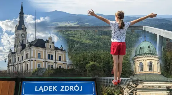 Dziewczynka z rozłożonymi rękami podziwia panoramę Lądka Zdroju z pięknymi budynkami i górami w tle.