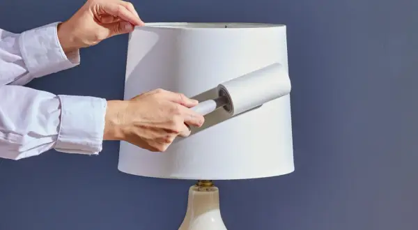 Hands using a lint roller to clean a white lampshade, demonstrating how to clean lamp shades effectively.