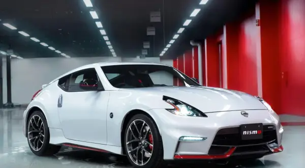Biały Nissan 370Z Nismo z czerwonymi akcentami, lśniący w garażu z czerwonymi ścianami i rzędem świateł na suficie.