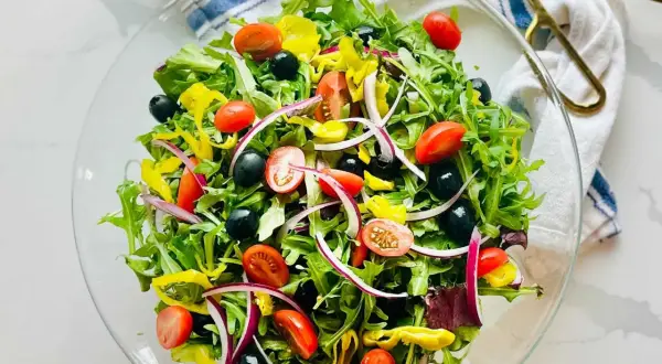 En fräsch italiensk sallad i en glasskål, fylld med gröna blad, körsbärstomater, svarta oliver, rödlök och gul paprika.