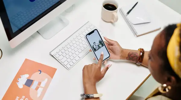 Une développeuse consulte son blog sur son téléphone, entourée d'un ordinateur, d'une tasse de café et de documents de conception.