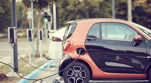 Due **utilitarie elettriche** arancioni e nere, una bianca, sono in carica. Stazioni di ricarica con cavi collegati.