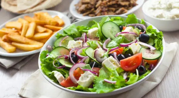 Świeża sałatka z sałatą, pomidorami, ogórkiem, fetą i oliwkami – idealne co do sałatki gyros. Obok frytki i mięso gyros.