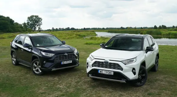 Dwa nowe modele Toyota RAV4 2025, czarny i biały, zaparkowane na trawie nad jeziorem.