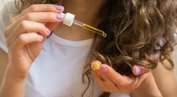 Hände mit lila und gelbem Nagellack tragen Rizinusöl (castor oil) auf lockiges Haar auf.