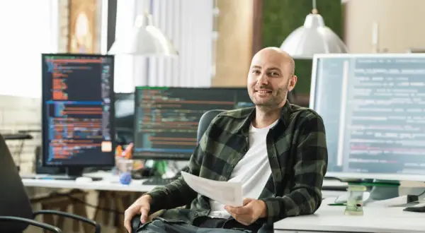 Ein Mann mit Glatze, der ein Dokument hält, sitzt vor mehreren Computermonitoren mit Code. Er könnte einen Techniker im Fernstudium bei der IHK absolvieren.