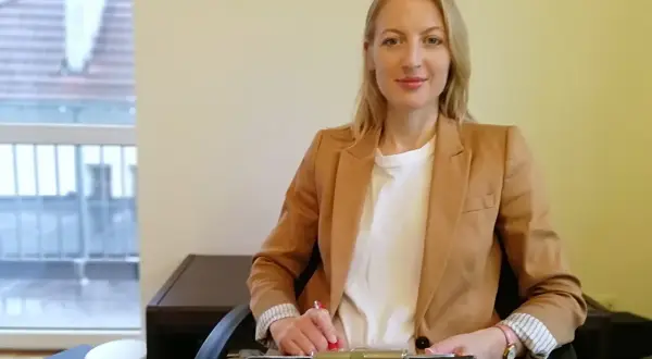 Kobieta w beżowej marynarce siedzi przy biurku i pisze.