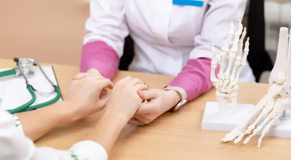 Arzt hält Patientin die Hand. Modelle von Fuß und Hand liegen auf dem Tisch. Hoffnung auf eine Wunderwaffe gegen Arthrose.