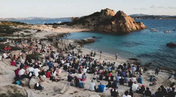 Concerto sulla spiaggia in Sardegna, con un pubblico seduto tra le rocce che ascolta un cantante sardo.