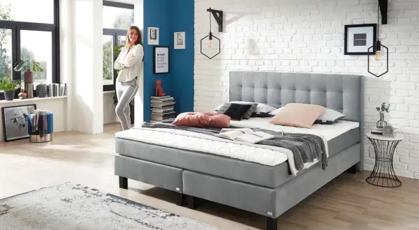 Modernes Boxspringbett in Grau, ideal für erholsamen Schlaf bei Bandscheibenvorfall. Eine Frau lehnt lässig daneben.