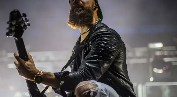 Charyzmatyczny gitarzysta z brodą podczas koncertu Bullet for My Valentine, w skórzanej kurtce i podartych spodniach, gra na gitarze.