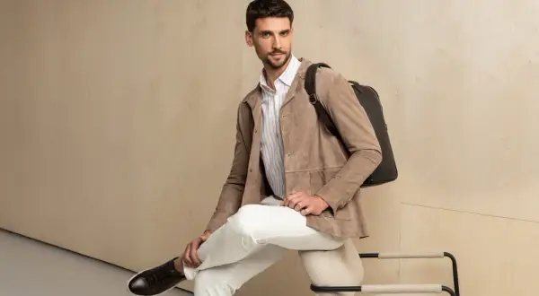 Elegancka stylizacja męska: beżowa kurtka z zamszu, białe spodnie, koszula w paski i czarny plecak.