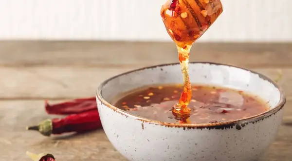En träsked droppar gyllene chili honung ner i en skål. Torkade chilifrukter ligger bredvid.