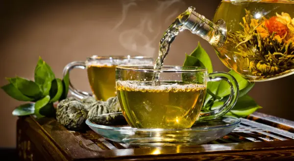 Gorąca herbata parzy się w szklanej filiżance, a z dzbanka wlewa się aromatyczny napar. Obok leżą zielone kulki herbaty.