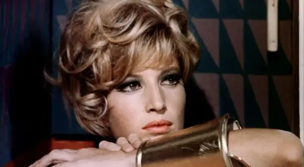Monica Vitti, con un bracciale dorato e un'espressione pensierosa, sfoggia un look audace con capelli biondi cotonati e trucco marcato.