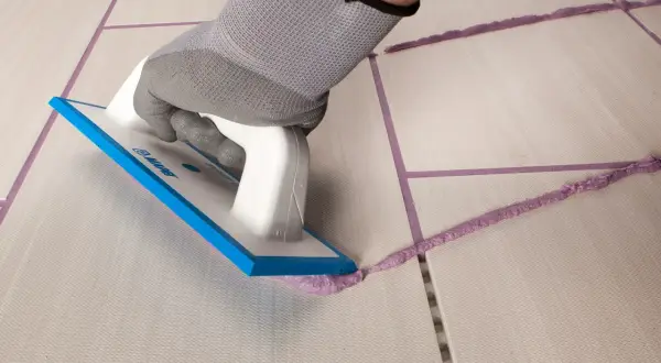 Mano con guanto grigio stende cemento e acqua senza sabbia con spatola su fughe di piastrelle.