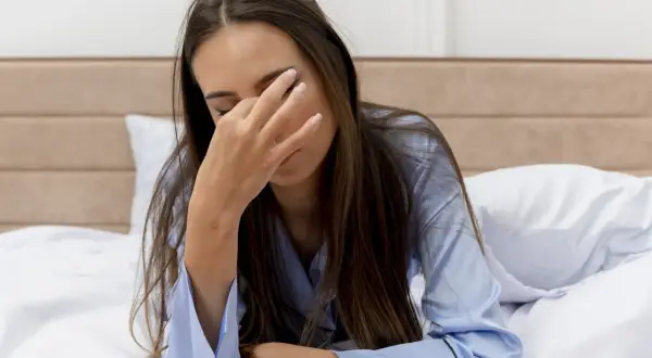 Een vrouw ligt moe in bed, haar hand voor haar ogen. Ze draagt een blauwe pyjama.