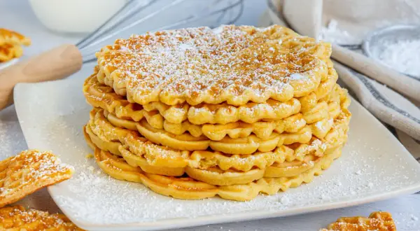 Stapel goldbrauner Waffeln ohne Backpulver, bestäubt mit Puderzucker. Daneben liegen weitere Waffeln.