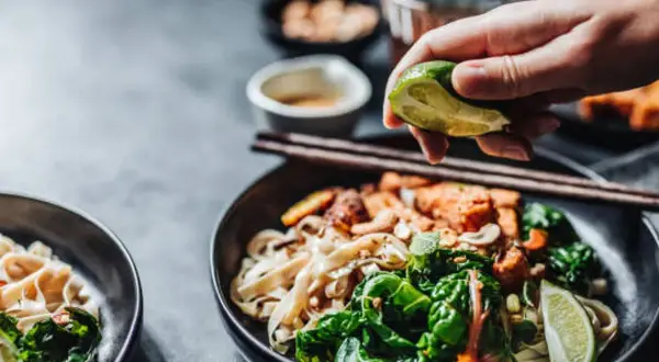 En hand pressar lime över en skål med nudlar och grönsaker, perfekt vin till asiatisk mat.