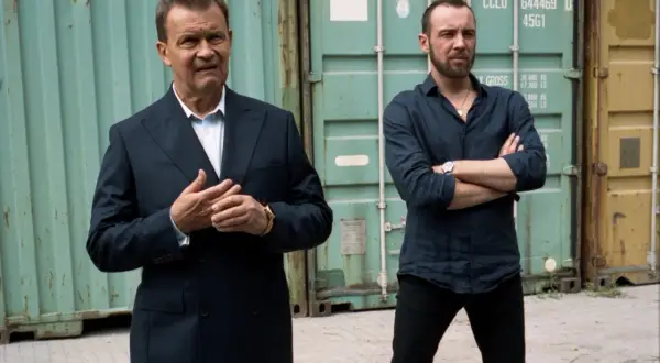 Dwóch mężczyzn, jeden w garniturze, drugi w koszuli, stoi przed kontenerami. Scena jak z filmu "Jak zostałem gangsterem. Cytaty" o życiu w półświatku.