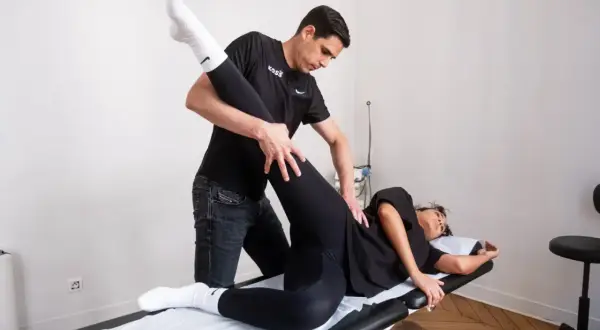 Un kinésithérapeute guide un exercice kiné hanche, soulevant la jambe d'une patiente allongée sur une table de massage.